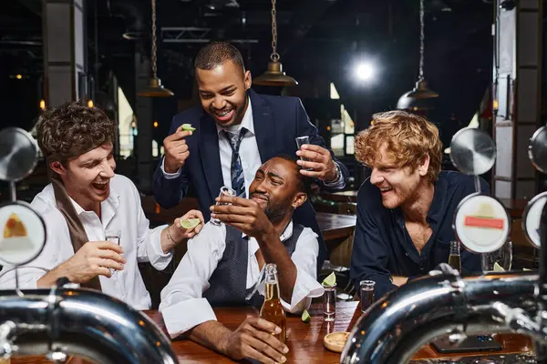 Группа из четырех счастливых и пьяных многонациональных друзей в формальной одежде, пьющих текилу в баре — стоковое фото