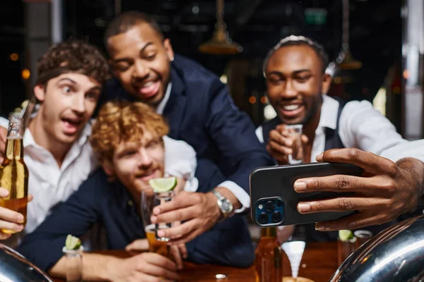 Cheerful multiethnic colleagues in formal wear taking selfie on smartphone in bar after work — Stock Photo