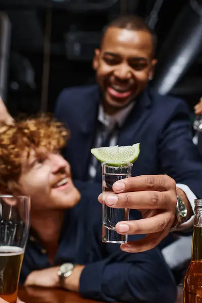 Focus sur tequila shot avec du citron vert tranché sur le dessus, homme afro-américain tenant boisson près d'un ami dans le bar — Photo de stock