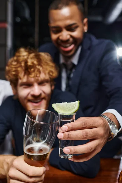 Dos colegas interracial tintineo con vasos después del trabajo en el bar, tequila y vaso de cerveza - foto de stock