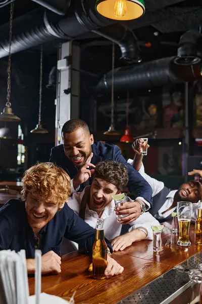 Drunk and funny interracial men in formal wear laughing while drinking tequila and beer in bar — Stock Photo