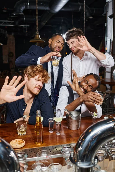 Cuatro amigos borrachos interracial cubriendo caras de luz brillante, tequila, cerveza y whisky - foto de stock