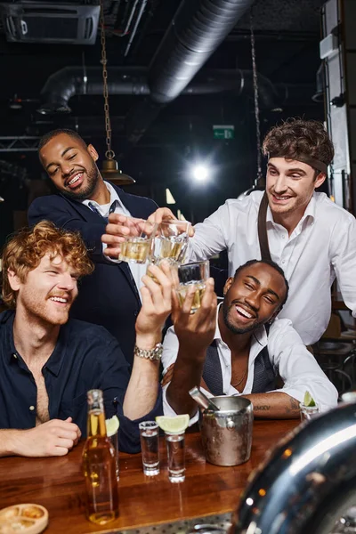 Quatre amis multiculturels heureux griller avec des verres de whisky près de tirs de tequila et de bière dans le bar — Photo de stock