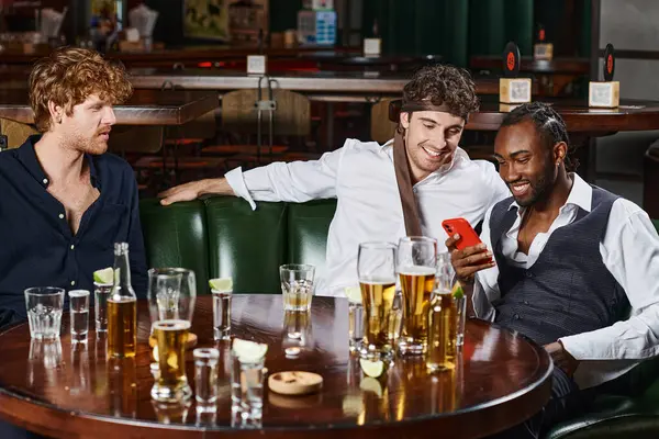 Hombre afroamericano feliz usando teléfono inteligente y pasar tiempo con colegas después del trabajo en el bar - foto de stock