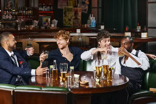 Vier fröhliche multikulturelle Freunde trinken Whiskey in der Nähe von Tequila-Shots und Bier auf dem Tisch in der Bar — Stockfoto