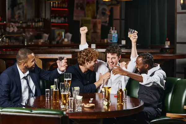 Drunk man raising hands with glass of whiskey near multiethnic friends in bar, after work fun — Stock Photo