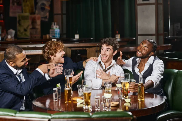 Hombres multiétnicos felices apuntando a un amigo en el bar, amigos varones durante la despedida de soltero en el bar - foto de stock
