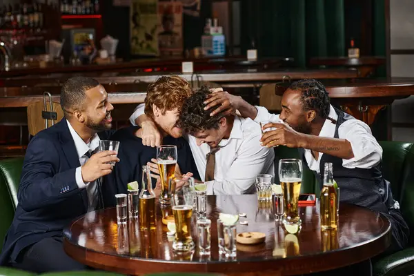 Amigos masculinos multiétnicos felices abrazándose durante la despedida de soltero en bar, tequila, whisky y cerveza - foto de stock