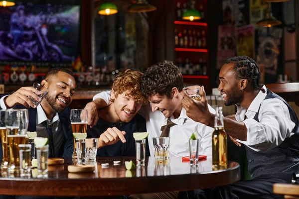 Happy and drunk multiethnic friends hugging during bachelor party in bar, tequila, whiskey and beer — Stock Photo