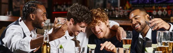 Happy and drunk multiethnic friends hugging during bachelor party in bar, alcohol drinks banner — Stock Photo