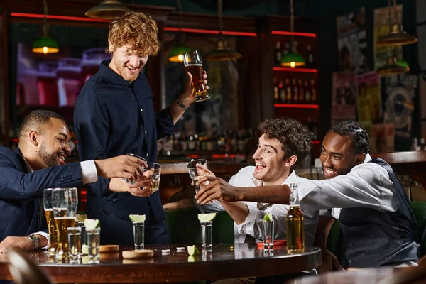 Happy and drunk multiethnic colleagues toasting with glasses of whiskey during bachelor party in bar — Stock Photo