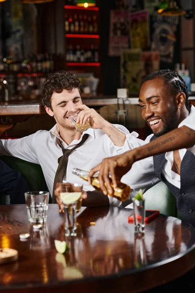 Happy drunk man drinking whiskey near cheerful african american friend pouring beer in glass — Stock Photo