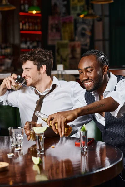 Homem americano africano alegre derramando cerveja em vidro perto de amigo sorrindo e bebendo uísque — Fotografia de Stock