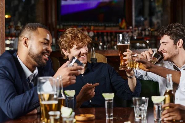 Feliz pelirrojo hombre apuntando a amigo mientras toma bebidas después del trabajo en el bar, amigos interracial - foto de stock