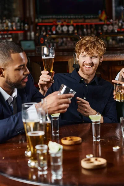 Cheerful redhead man pointing at friend while having drinks after work in bar, interracial friends — Stock Photo