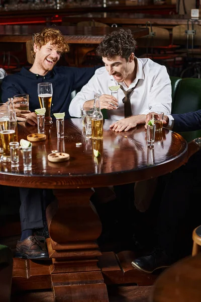 Drunk man trying to bite lime on top of tequila shot near cheerful friend, spending time after work — Stock Photo