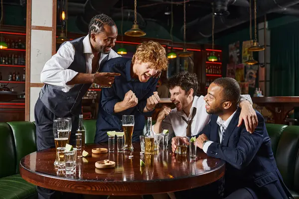 Drunk interracial friends laughing and drinking alcohol drinks while relaxing after work in bar — Stock Photo