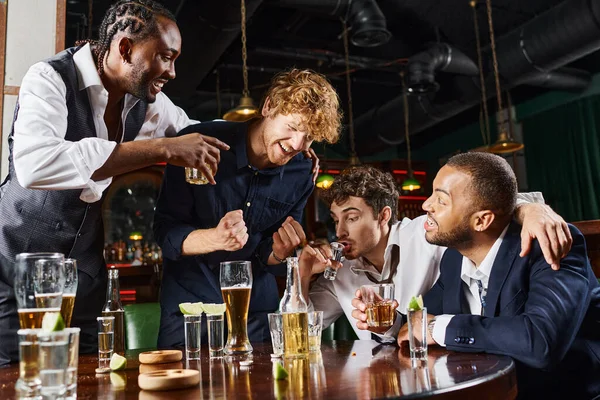 Happy interracial friends laughing and drinking alcohol drinks while relaxing after work in bar — Stock Photo