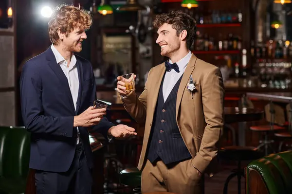 Happy groom in suit with boutonniere and best man holding glasses of whiskey during bachelor party — Stock Photo