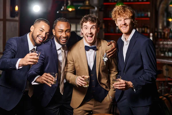 Marié heureux debout avec des amis interracial et tenant des verres de whisky pendant l'enterrement de vie de garçon — Photo de stock