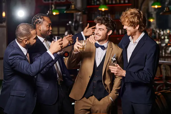 Amici felici in abiti formali congratularsi con lo sposo nel bar, uomini interrazziali che tengono bicchieri di whisky — Foto stock