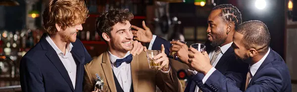 Amis heureux en tenue formelle félicitant marié dans le bar, hommes interracial tenant bannière de whisky — Photo de stock