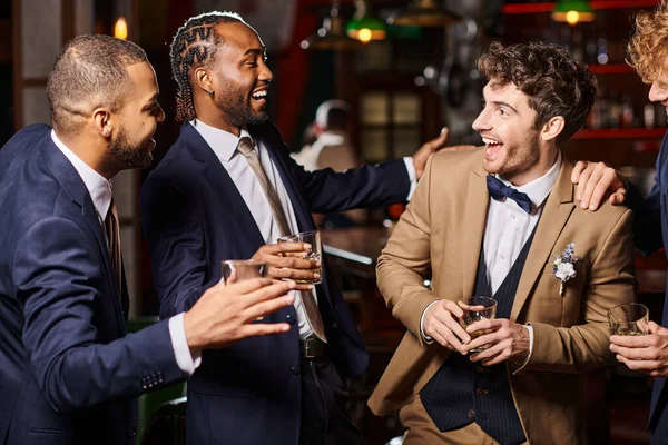 Feliz interracial amigos en formal desgaste felicitando novio en bar, hombres sosteniendo vasos de whisky - foto de stock