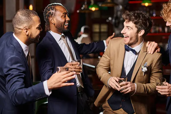 Happy interracial friends in formal wear congratulating groom in bar, men holding glasses of whiskey — Stock Photo