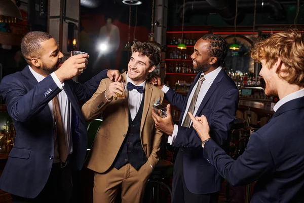 Feliz interracial novios en formal desgaste felicitando amigo en el bar, los hombres con vasos de whisky - foto de stock