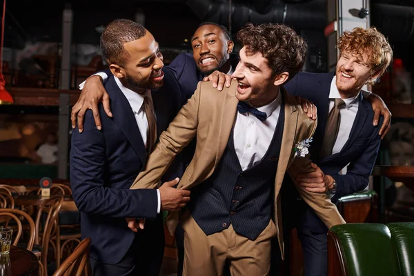 Enterrement de vie de garçon, heureux interracial hommes félicitant ami dans le bar, meilleurs hommes et marié en costumes — Photo de stock