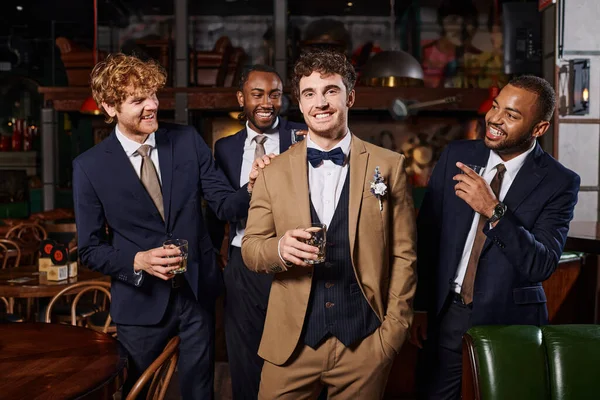 Холостяцька вечірка, щасливі міжрасові кращі чоловіки, дивлячись на нареченого в костюмі, що стоїть з віскі в барі — стокове фото