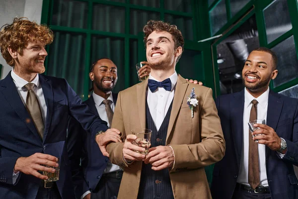 Despedida de soltero, alegre interracial hombres felicitando amigo en bar, mejores hombres y novio en trajes - foto de stock