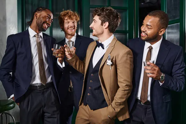Despedida de soltero, multiétnicos mejores hombres tostando vasos de whisky con elegante novio en el bar - foto de stock