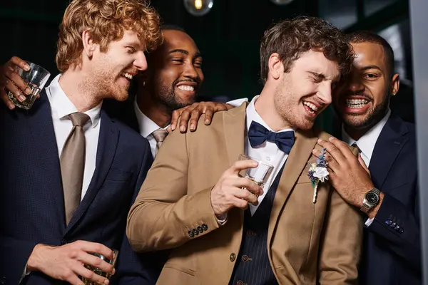 Despedida de soltero, excitados mejores hombres multiétnicos y novio riendo en el bar, sosteniendo vasos de whisky - foto de stock