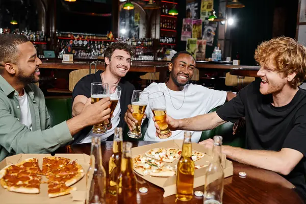 Noche fuera, feliz interracial hombres tintineo vasos de cerveza cerca de pizza en bar, amistad masculina - foto de stock