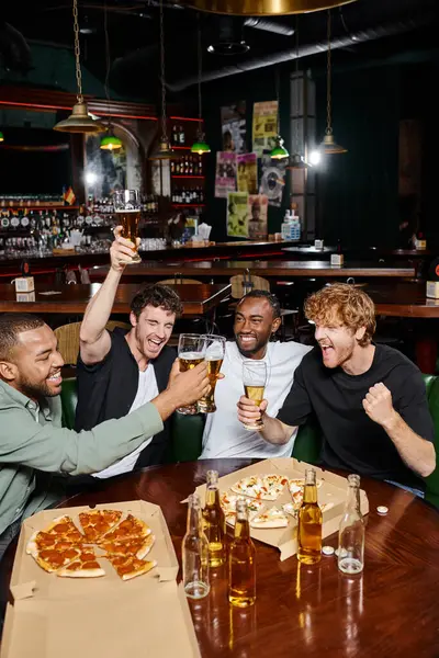 Ночь на улице, возбужденные межрасовые мужчины звон стаканов пива возле пиццы в баре, мужская дружба — стоковое фото
