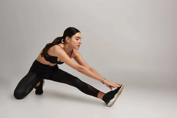 Mulher atraente no desgaste ativo preto com rastreador de fitness no pulso esticando a perna no fundo cinza — Fotografia de Stock