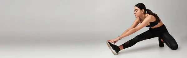Atraente jovem esportista em preto ativo desgaste alongamento perna no fundo cinza, banner — Fotografia de Stock