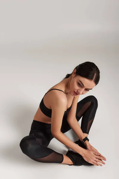 Sportiva flessibile in abito attivo nero con fitness tracker al polso seduta su sfondo grigio — Foto stock