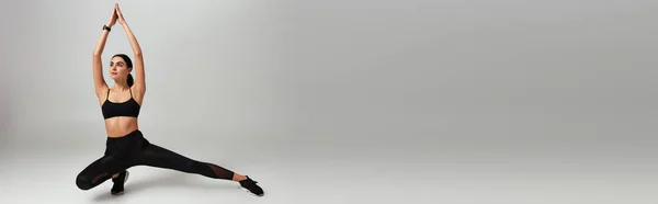 Молода спортсменка в чорному спортивному одязі робить легені руками над головою на сірому фоні, банер — стокове фото