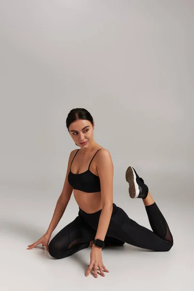 Flexible junge Sportlerin in schwarzen Leggings und bauchfreiem Top, die Beine vor grauem Hintergrund streckt — Stockfoto