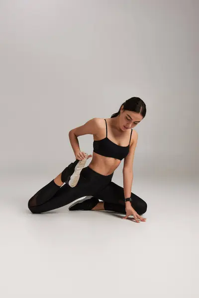 Deportiva flexible y delgada en desgaste activo negro y zapatillas de deporte que estiran las piernas sobre fondo gris - foto de stock