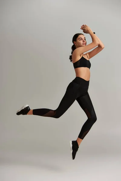 Young fit woman in black leggings, crop top and sneakers jumping on grey background, levitation — Stock Photo