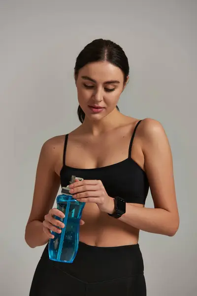 Mujer en forma de leggins y la parte superior de la cosecha celebración de la botella con agua, mientras que de pie sobre fondo gris - foto de stock