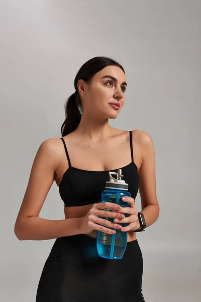 Retrato de mulher sonhadora em sportswear segurando garrafa com água enquanto em pé sobre fundo cinza — Fotografia de Stock