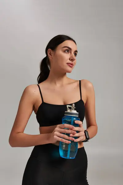 Portrait of dreamy woman in sportswear holding bottle with water while standing on grey background — Stock Photo