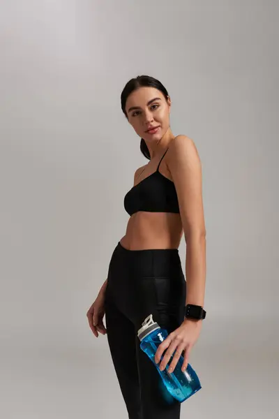 Portrait of sporty woman in active wear holding bottle with water while standing on grey background — Stock Photo