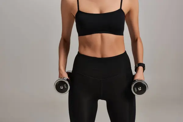 Vista ritagliata di forte sportiva in nero attivo usura esercizio con manubri su sfondo grigio — Foto stock