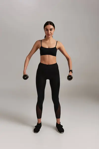 Longitud completa de la deportista fuerte en el ejercicio de desgaste activo negro con mancuernas sobre fondo gris - foto de stock