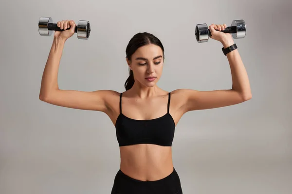 Jovem mulher motivada e esportiva em desgaste ativo preto trabalhando com halteres em fundo cinza — Fotografia de Stock